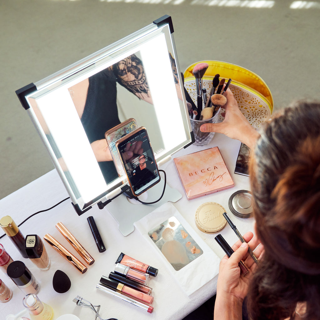 A birds-eye view of a woman using a TML Meira makeup vanity, her phone mounted to the mirror. Makeup scattered on the table.