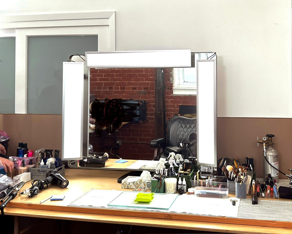 A mirror its atop a table with makeup and tools. On the top and two sides of the mirror are TML light panels.