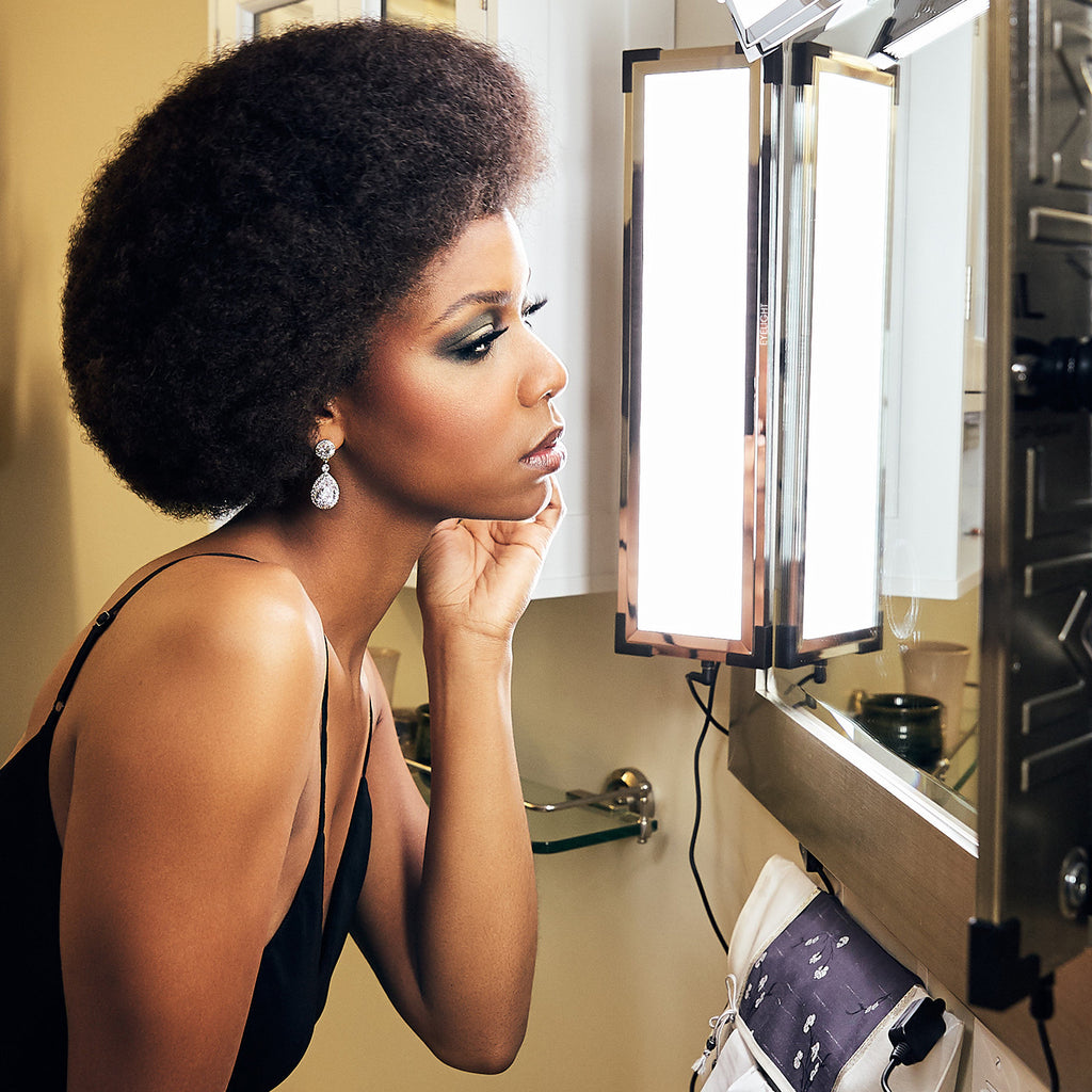 A woman looks at her reflection in a mirror, illuminated by The Makeup Light Eyelights