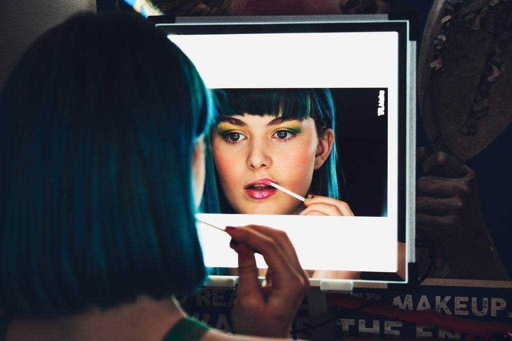 A teenage woman with blue hair and bags applies lipgloss in the light of her TML Meira