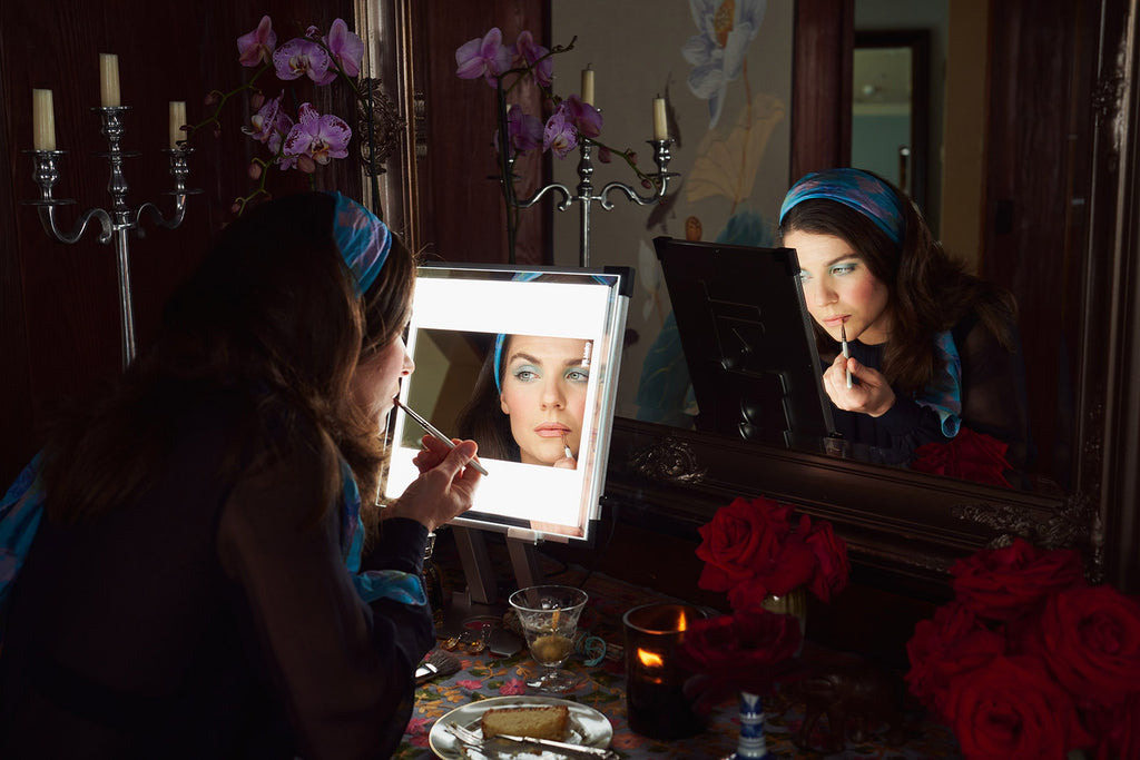 A young woman wearing a headband applies makeup using her TML Meira tabletop vanity. A wall mirror behind catches her reflection, illuminated by the Meira.