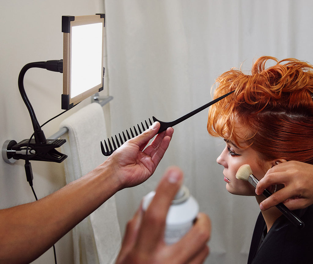A Key Light clamped onto a towel rod on the left - model with makeup artist hands and hair artist hands on the right
