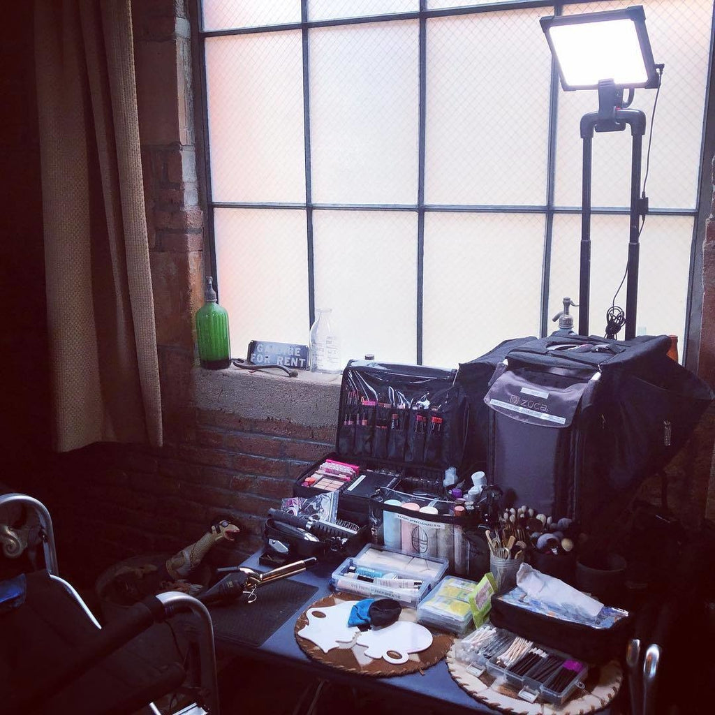 A TML LED Key Light affixed atop the handle of a luggage bag on a clamp. Makeup products and tools on the table below.