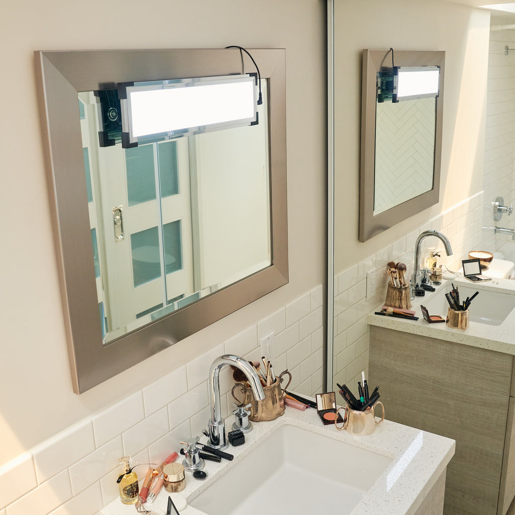 A single TML LED Eyelight affixed flush to a bathroom mirror. Makeup products and tools surround the edges of the sink below.